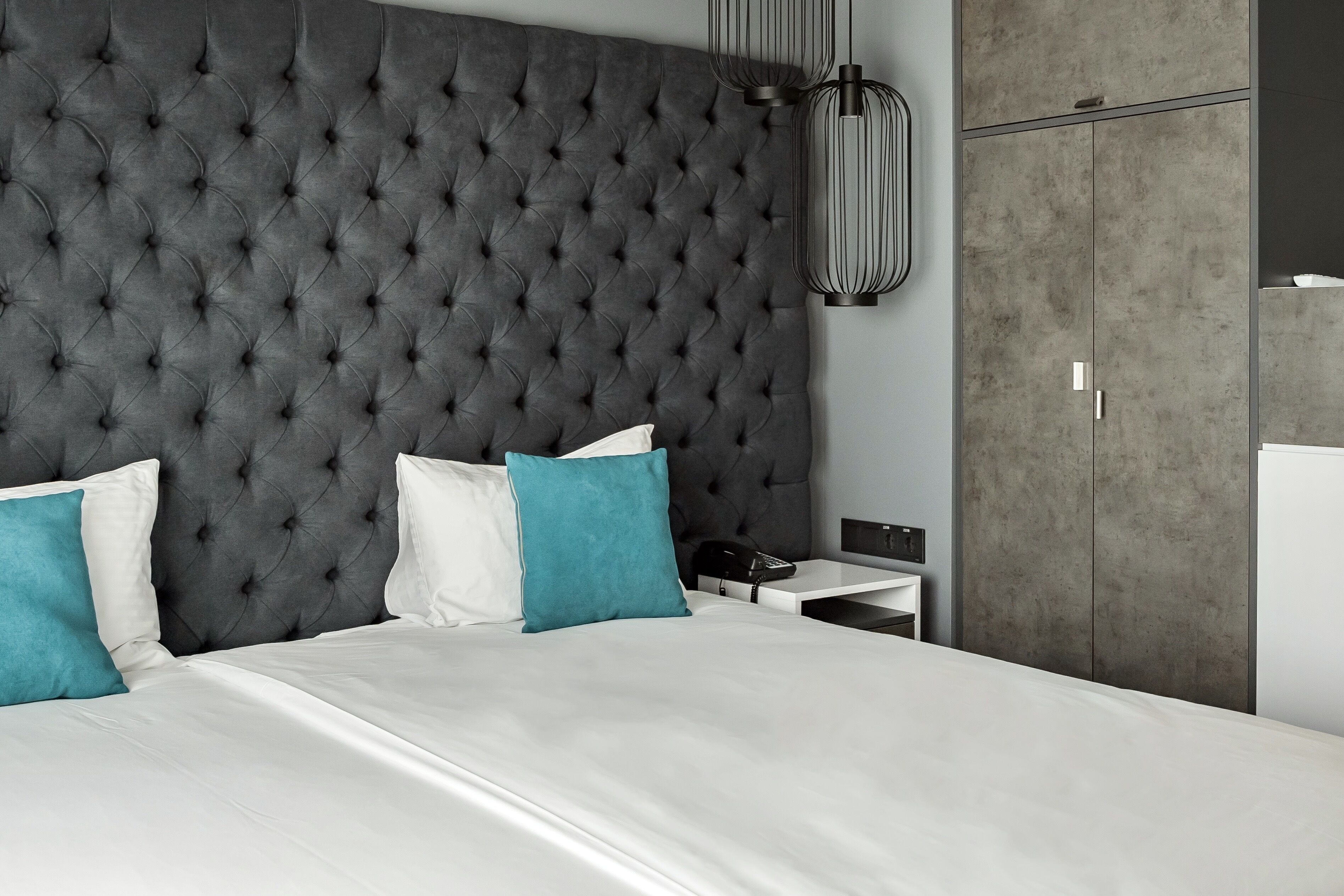 A modern bedroom with a tufted headboard, a white bed, teal and white pillows, a bedside table with a lamp, and a wardrobe.