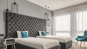 Una habitación de hotel moderna con dos camas, un cabecero gris con capitonado, una silla azul y lámparas colgantes.