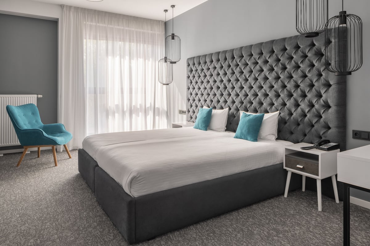 Una habitación de hotel moderna con una cama grande, un cabecero capitonado, un sillón verde azulado y un escritorio con teléfono.