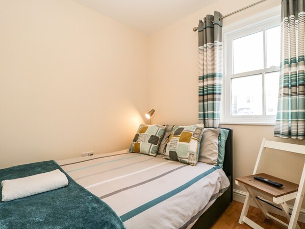 Een netjes opgemaakt bed met een gestreept dekbed en kussens, een bedlampje, een klein houten tafeltje met een afstandsbediening, en een raam met gestreepte gordijnen.
