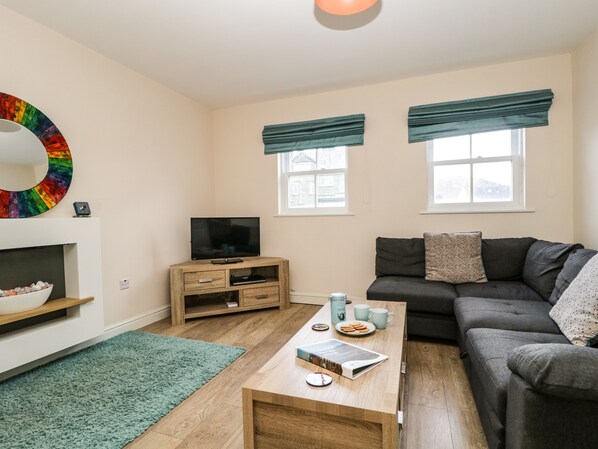 Een moderne woonkamer met een flatscreen televisie, een donkergrijze bank, een houten salontafel en een teal kleed.