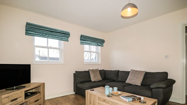 Ein Wohnzimmer mit einem dunkelgrauen Sofa, einem Holztisch und einem Fernsehschrank. Zwei Fenster mit blauen Vorhängen und eine Deckenleuchte sind vorhanden.