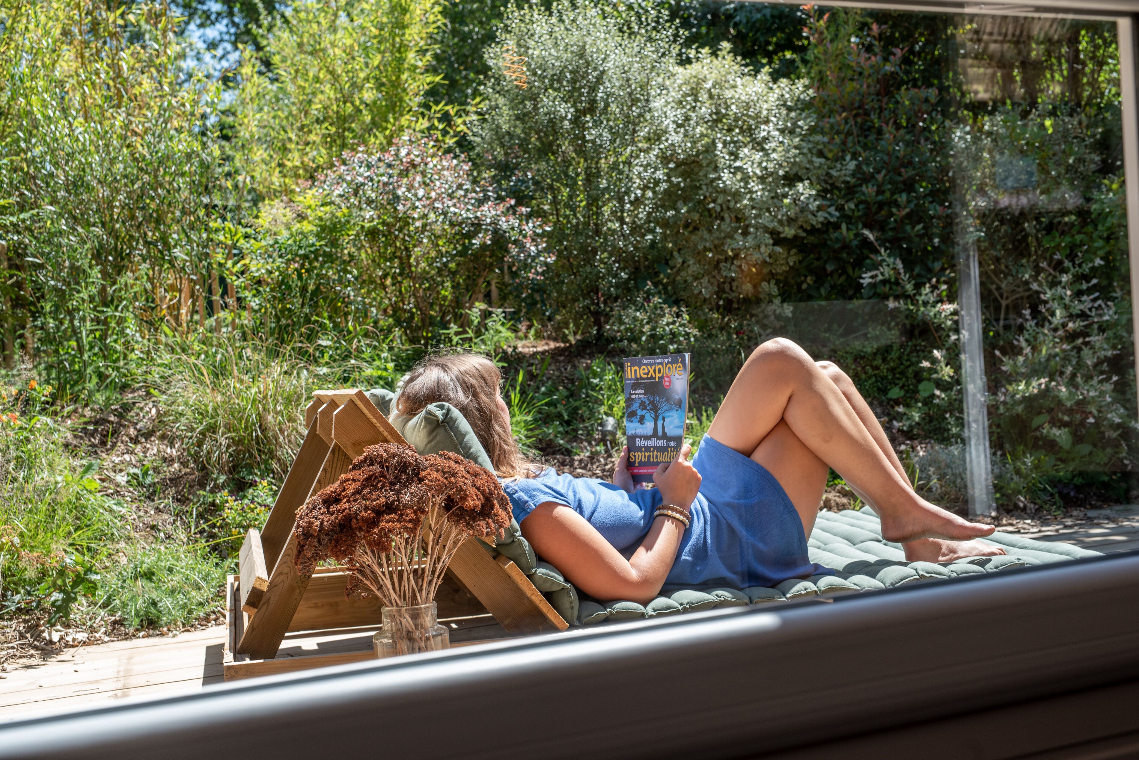 A person lying on a hammock in a garden with a book titled 'Revelations' and a wooden structure with dried flowers.