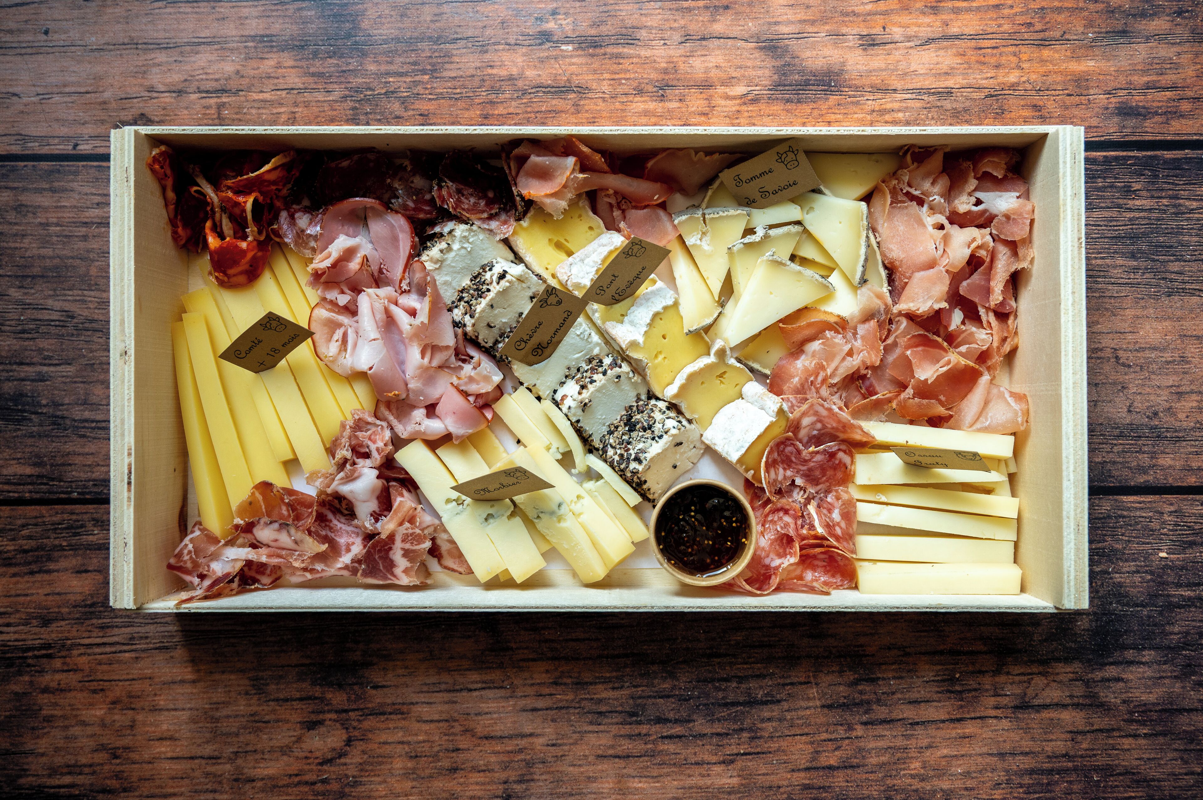 A wooden box containing various cheeses, cured meats, and a small bowl of black pepper.