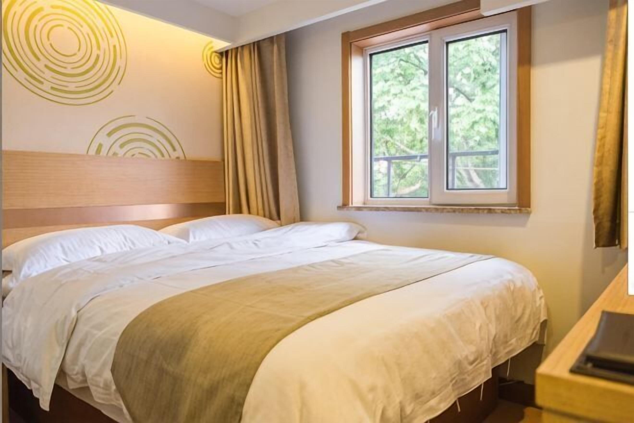 A neatly made bed with a headboard, a window with curtains, and a desk with a lamp.
