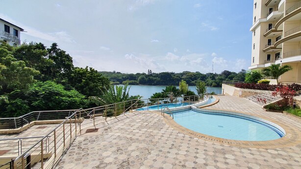 A swimming pool with a view of a lake, surrounded by greenery and a multi-story building. - The Beautiful 3Br All En-suite Apt (Mombasa)