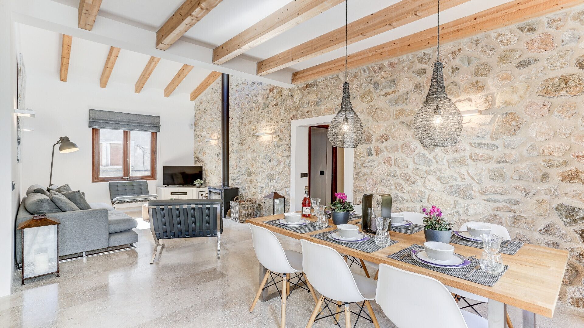 A modern living space with a stone wall, wooden beams, a dining area with white chairs, and a sofa with grey cushions.