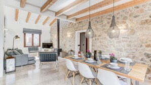 Un espace de vie moderne avec un mur en pierre, des poutres en bois, un coin repas avec des chaises blanches et un canapé avec des coussins gris.