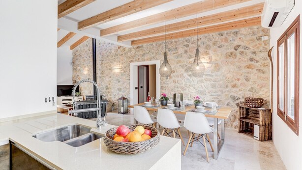 Une cuisine avec un mur en pierre, des poutres apparentes, un évier et un coin repas comprenant des chaises et une table dressée pour quatre personnes.