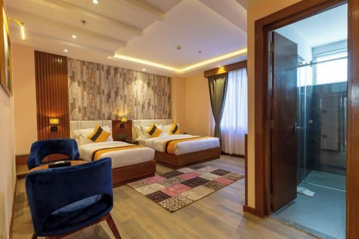 A hotel room with two beds, a blue chair, a wooden floor, a patterned rug, and a glass shower door.