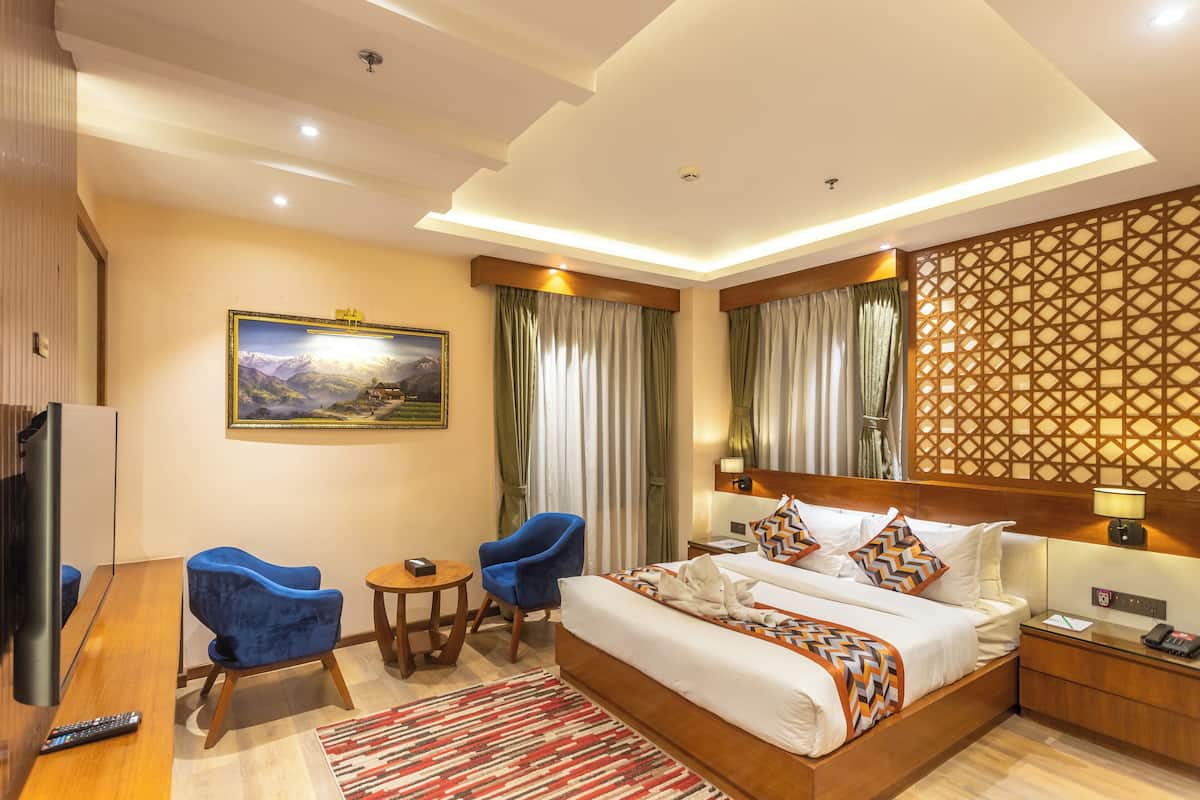 A hotel room with a large bed, two blue chairs, a small table, a TV, and a painting on the wall.