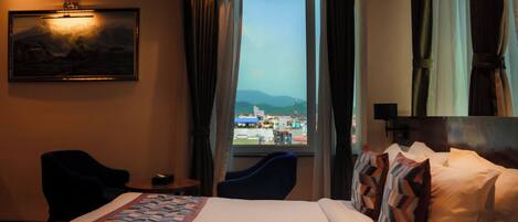 Habitación de hotel con cama, escritorio, silla, ventana con vista a la ciudad y un cuadro en la pared.