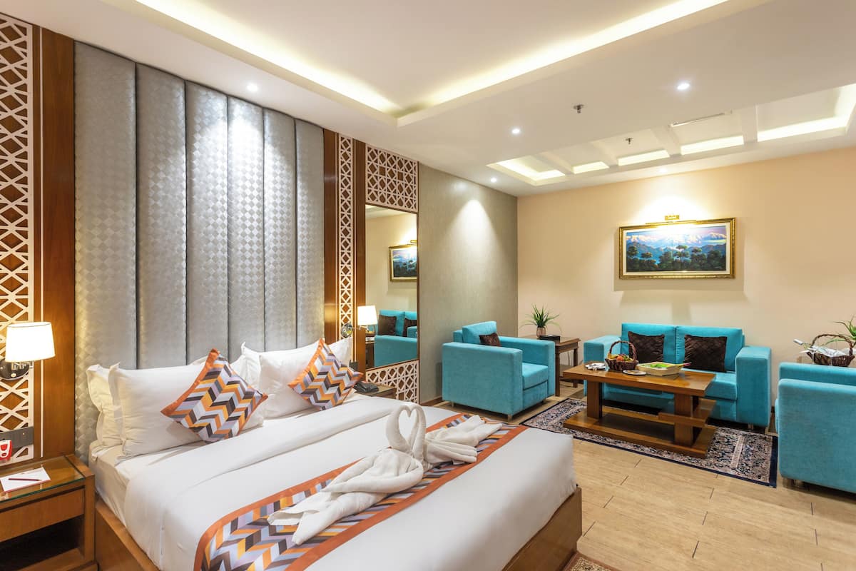 A hotel room with a large bed, two blue armchairs, a wooden coffee table, and a painting on the wall.