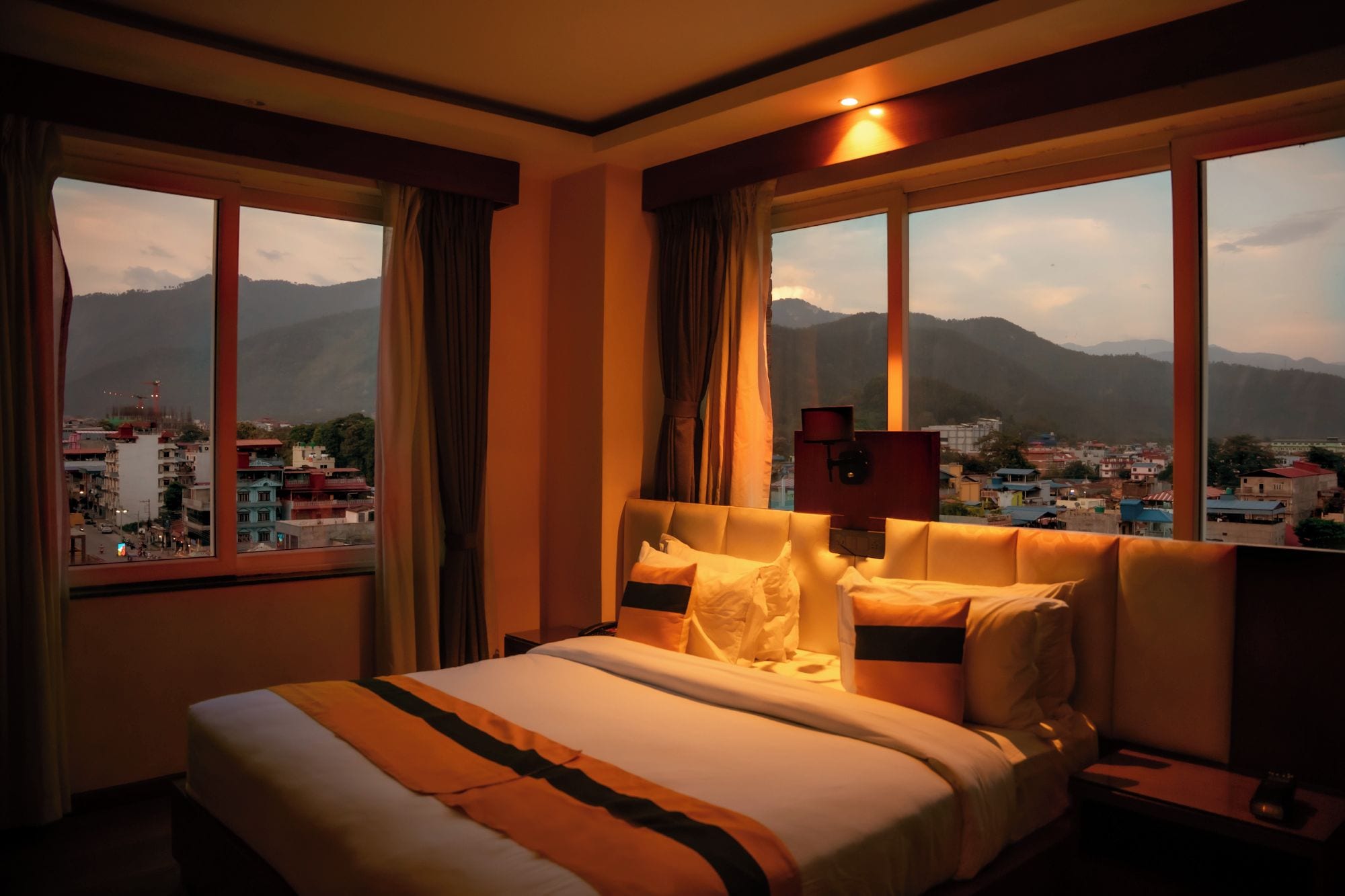Une chambre d’hôtel avec un lit, une table de chevet et une vue sur le paysage urbain et les montagnes, visible à travers de grandes fenêtres.
