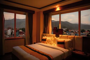 Une chambre d’hôtel avec un lit, une table de chevet et une vue sur le paysage urbain et les montagnes, visible à travers de grandes fenêtres.