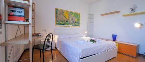 A bedroom with a bed, a chair, a desk with a chair, a bookshelf, and a painting on the wall.