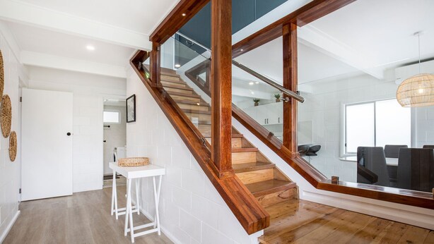 Un escalier avec des marches en bois et des rampes en verre, menant à un espace repas avec des chaises noires et un luminaire suspendu.
