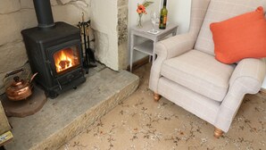 Un salon chaleureux avec une cheminée en pierre, un canapé agrémenté d’un coussin orange, une petite table avec une lampe et une bouteille, et une bouilloire posée sur le sol.