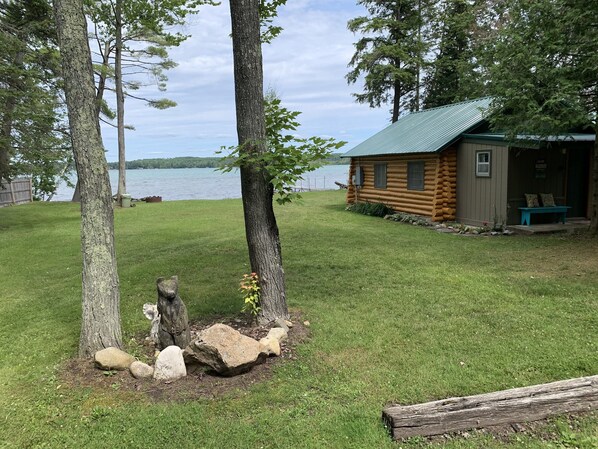 A cabin with a metal roof, surrounded by a grassy area with trees and a body of water in the background.