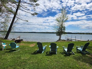 A lakeside view with four Adirondack chairs facing the water, a dock, and a grassy area with trees. - Waterfront log home on Elk Lake with dock and mooring, window A/C. Dog ok. (Williamsburg)