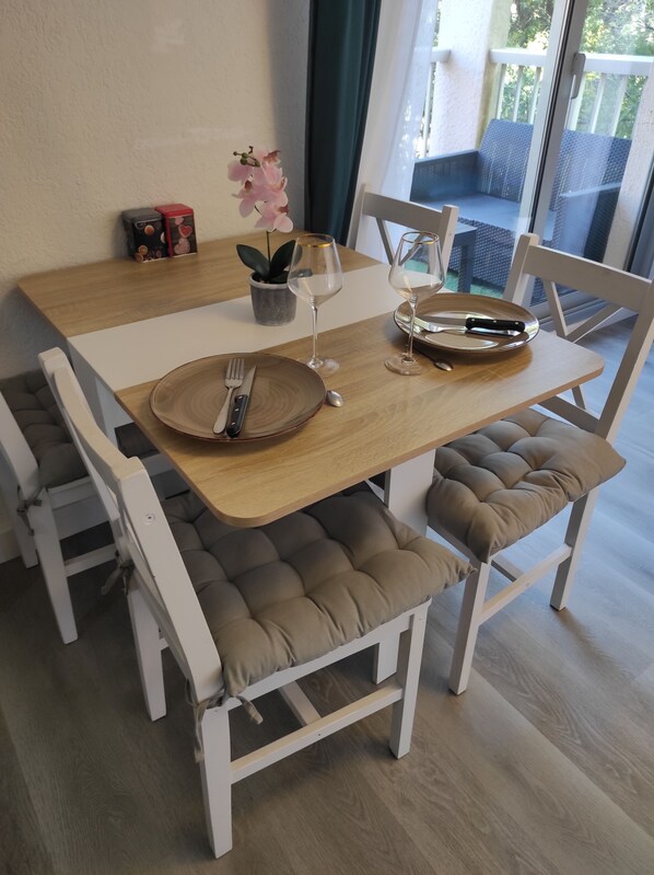 A dining area with a wooden table, white chairs with cushioned seats, a potted plant, and a glass of white wine.