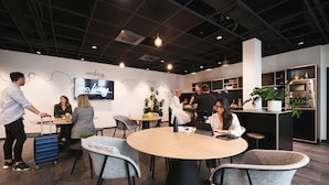 A modern interior with round tables and chairs, a kitchen area, and a TV displaying 'mo.living serviced apartments'. - MoLiving - Design Hotel & Apartments Düsseldorf-Neuss (Neuss)