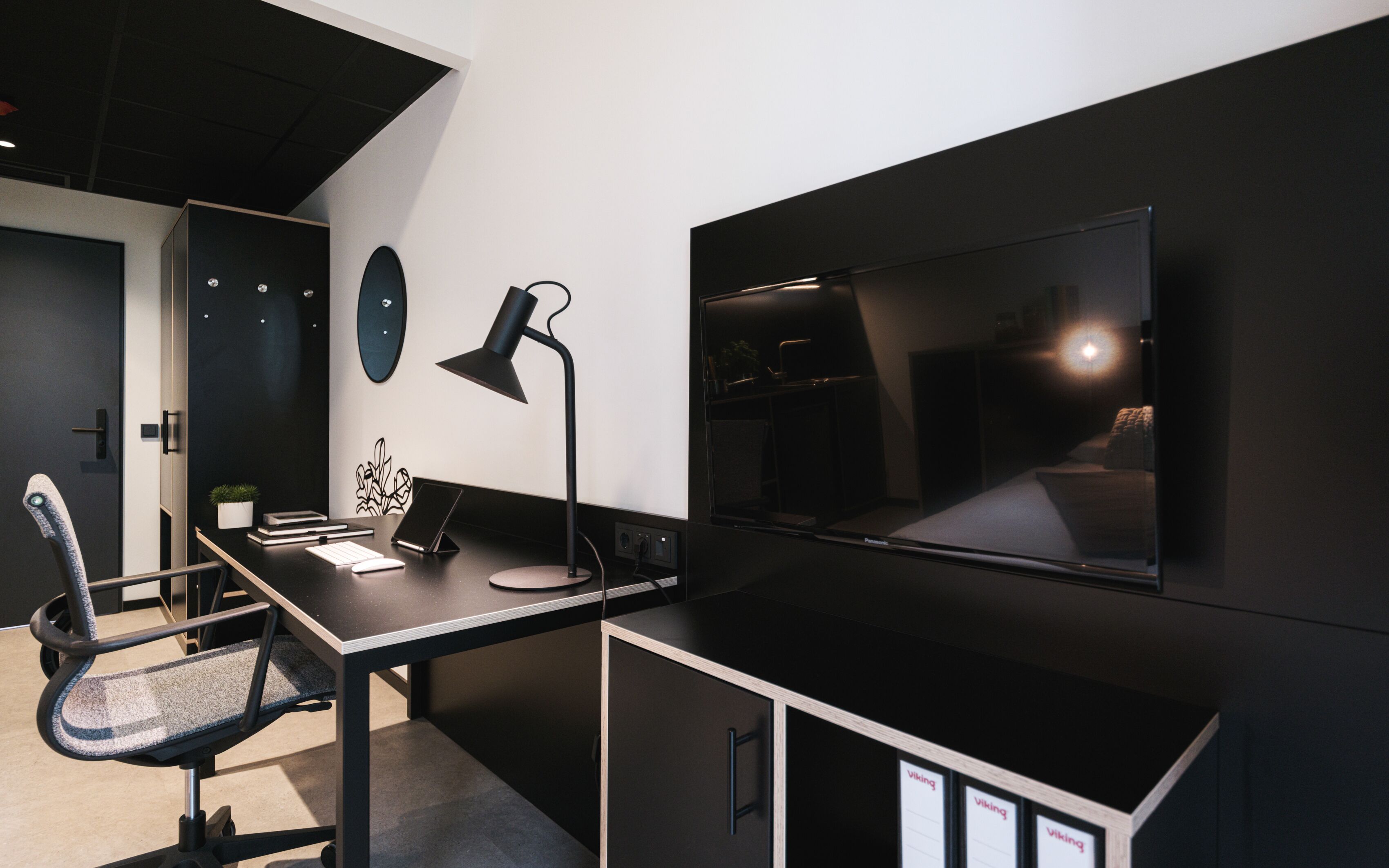 Un espace de bureau moderne comprenant un bureau, une chaise, un ordinateur, une lampe et un grand téléviseur à écran plat fixé au mur.