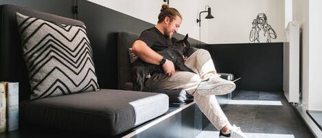 Una persona sentada en un sofá negro con un cojín estampado en rombos, sosteniendo un gato.
