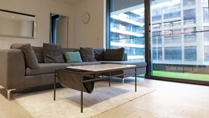 A modern living room with a grey sofa, a coffee table, and a large window offering a view of a building. - The Wardian (London)