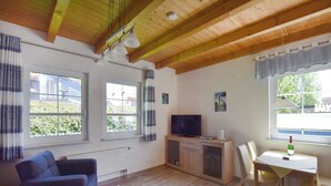 A cozy living room with a wooden ceiling, a television, a dining table with chairs, and large windows with curtains.