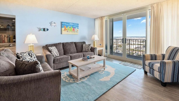 A living room with a sofa, armchair, and coffee table, a painting, and a view through a balcony. - Quay-Beautiful Beach Views in this 2 Bedroom Condo with Indoor Pool! (Ocean City)