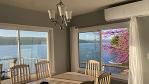 Een eetruimte met een tafel en stoelen, grote ramen met uitzicht op een meer en kersenbloesems, en een airconditioningsunit.