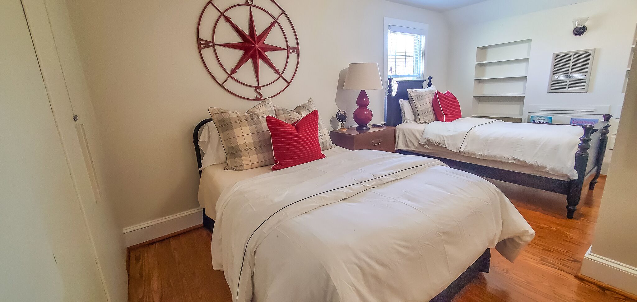 A bedroom with two beds, a red star decoration on the wall, a window with blinds, and a bedside table with a lamp.