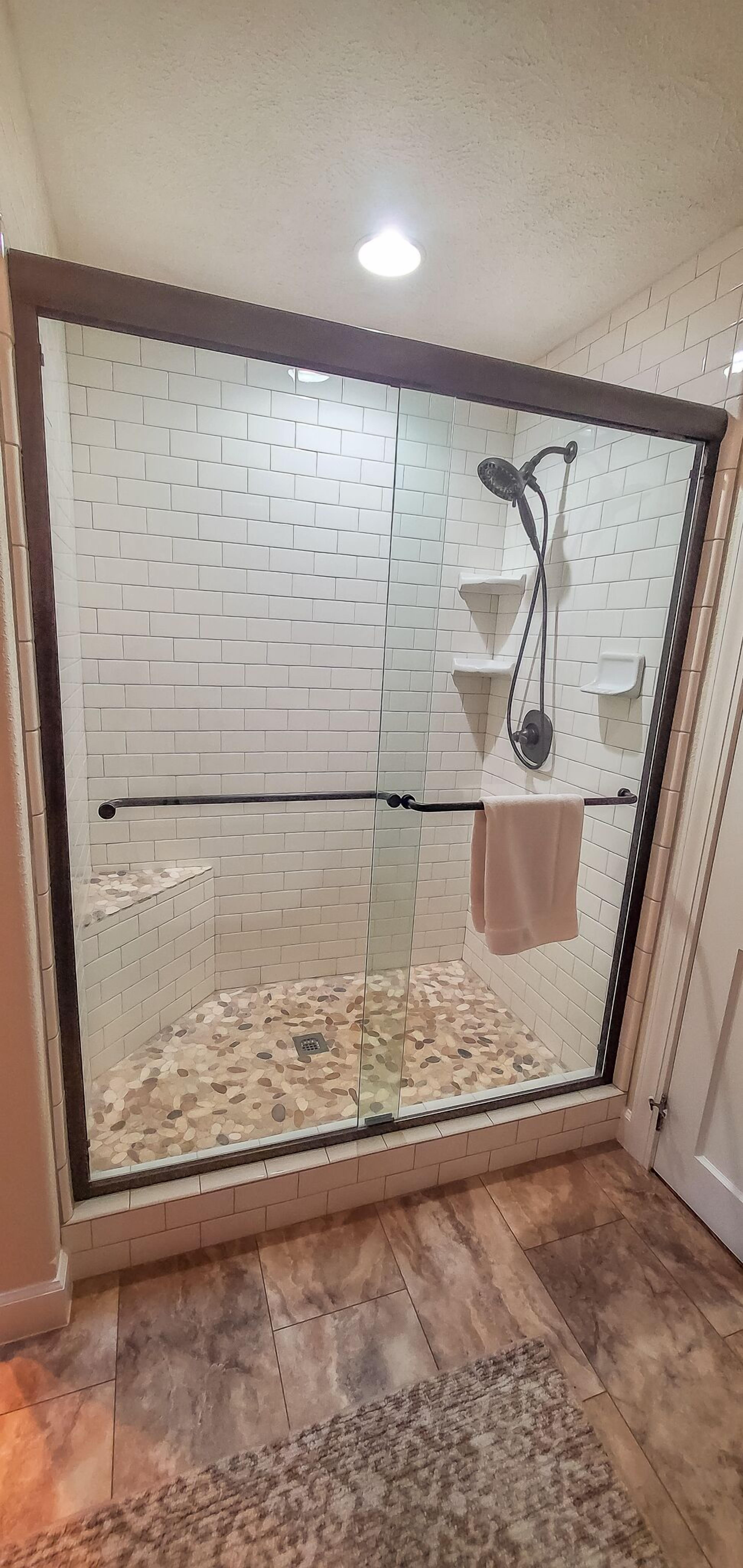 A glass-enclosed shower with white tiled walls and a pebble-patterned floor.