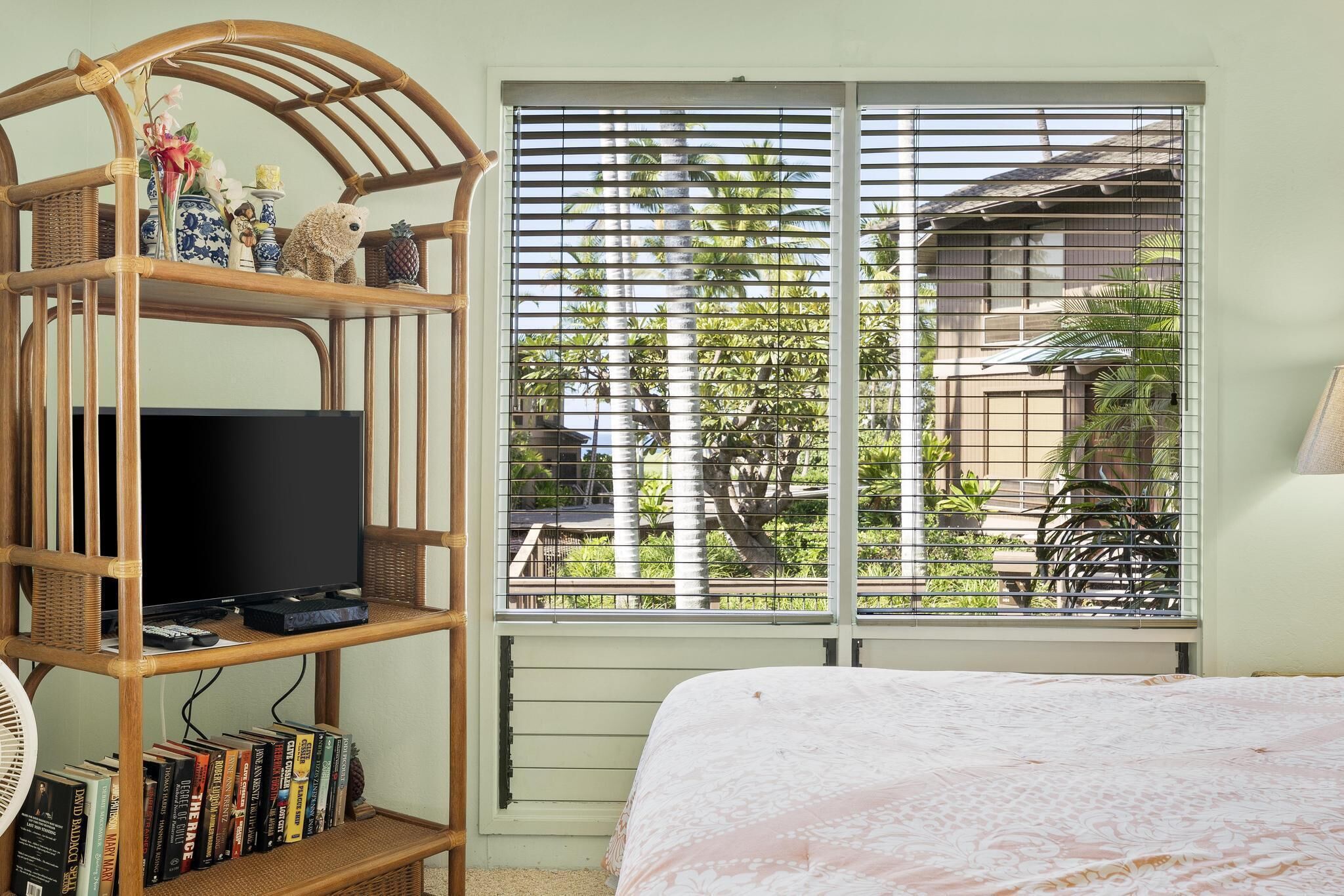 Ein Schlafzimmer mit einem Bett, einem Fernseher auf einem Holztisch, einem Fenster mit Jalousien und Blick ins Grüne.