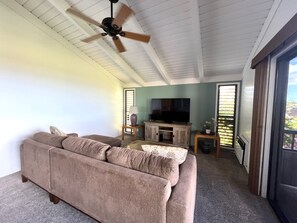 A living room with a beige sofa, a ceiling fan, a television on a wooden stand, and a view through a window with blinds.
