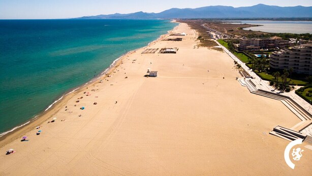 Une vue aérienne d’une plage avec une longue étendue de sable, quelques parasols et des bâtiments le long de la côte.