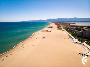 Vista aérea de una playa con una extensa franja de arena, algunos sombrillas y edificios a lo largo de la costa.