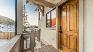 Eine Holztür mit einer Nummernschild, ein Balkon mit Holzgeländer und ein Blick auf eine verschneite Landschaft mit Bäumen und Bergen im Hintergrund.