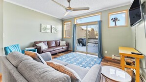 Un salon avec un canapé en cuir marron, un divan gris, un tapis à motifs bleus, un ventilateur de plafond et un téléviseur fixé au mur.
