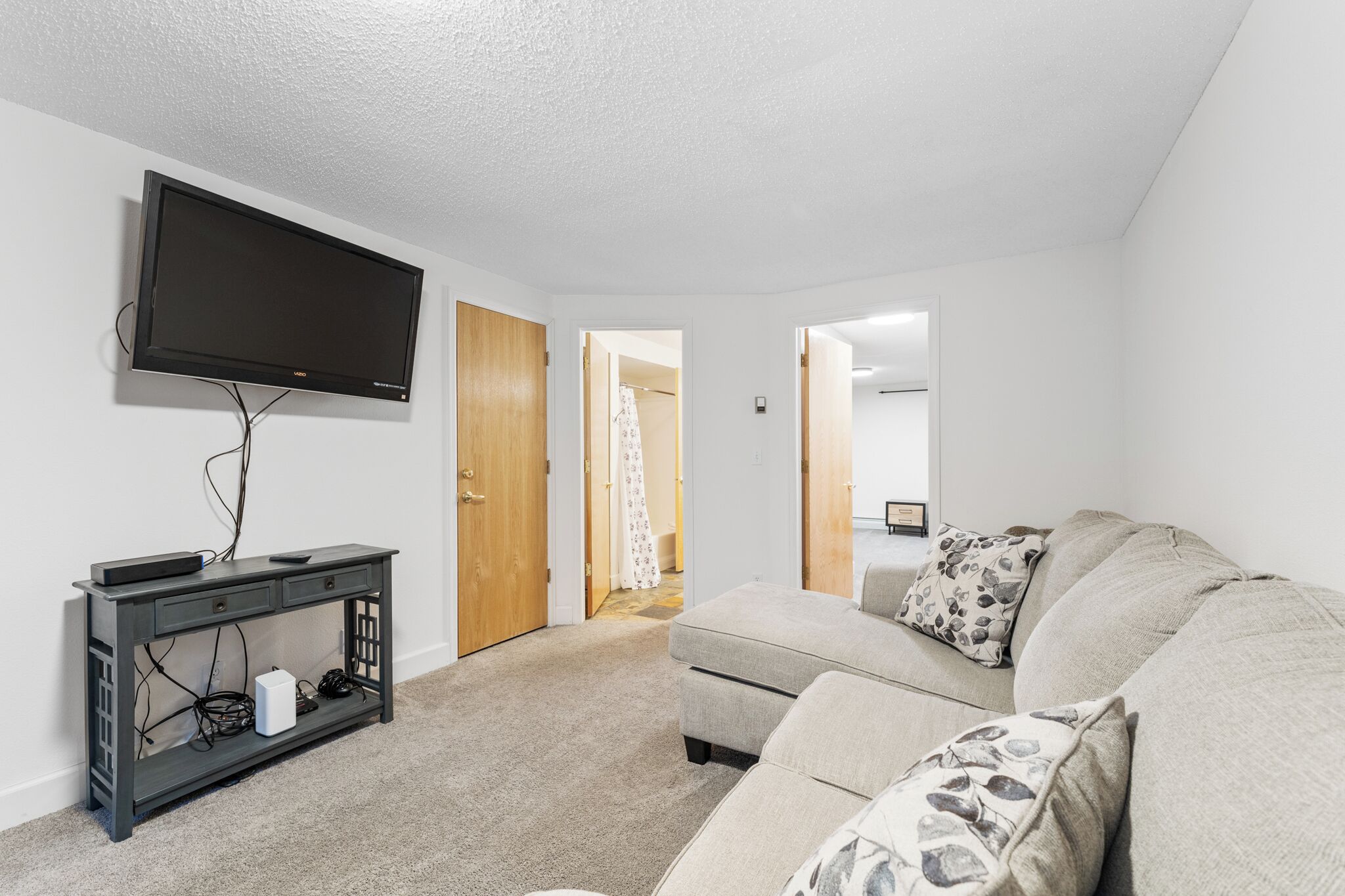 Ein Wohnzimmer mit einem an der Wand montierten Flachbildfernseher, einem dunklen Holzkonsole mit Schubladen, einem beige farbenen Sofa mit gemusterten Kissen und einem Teppichboden.