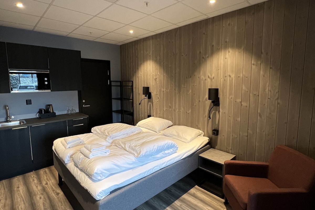 A modern bedroom with a large bed, a wooden accent wall, a small seating area, and a kitchenette.
