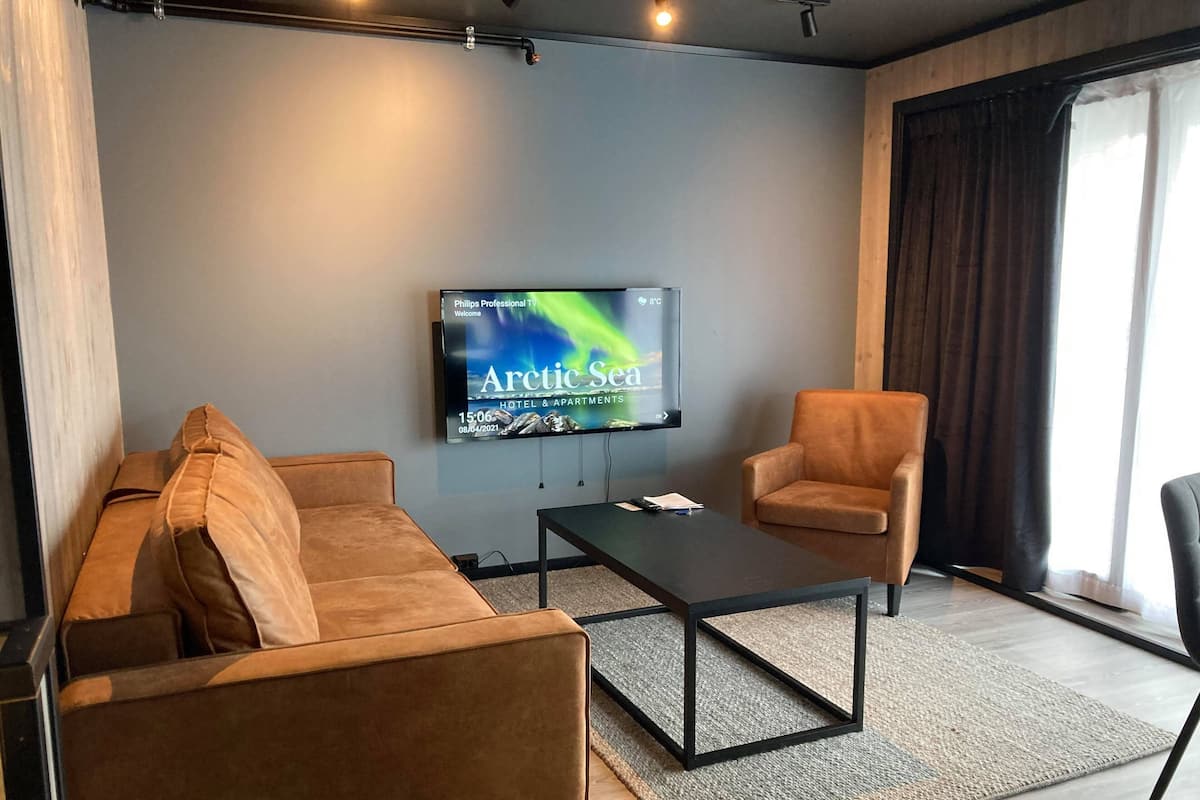 A modern hotel room with a large flat-screen TV displaying 'Arctic Sea' on the wall, a brown sofa, a matching armchair, and a small coffee table.