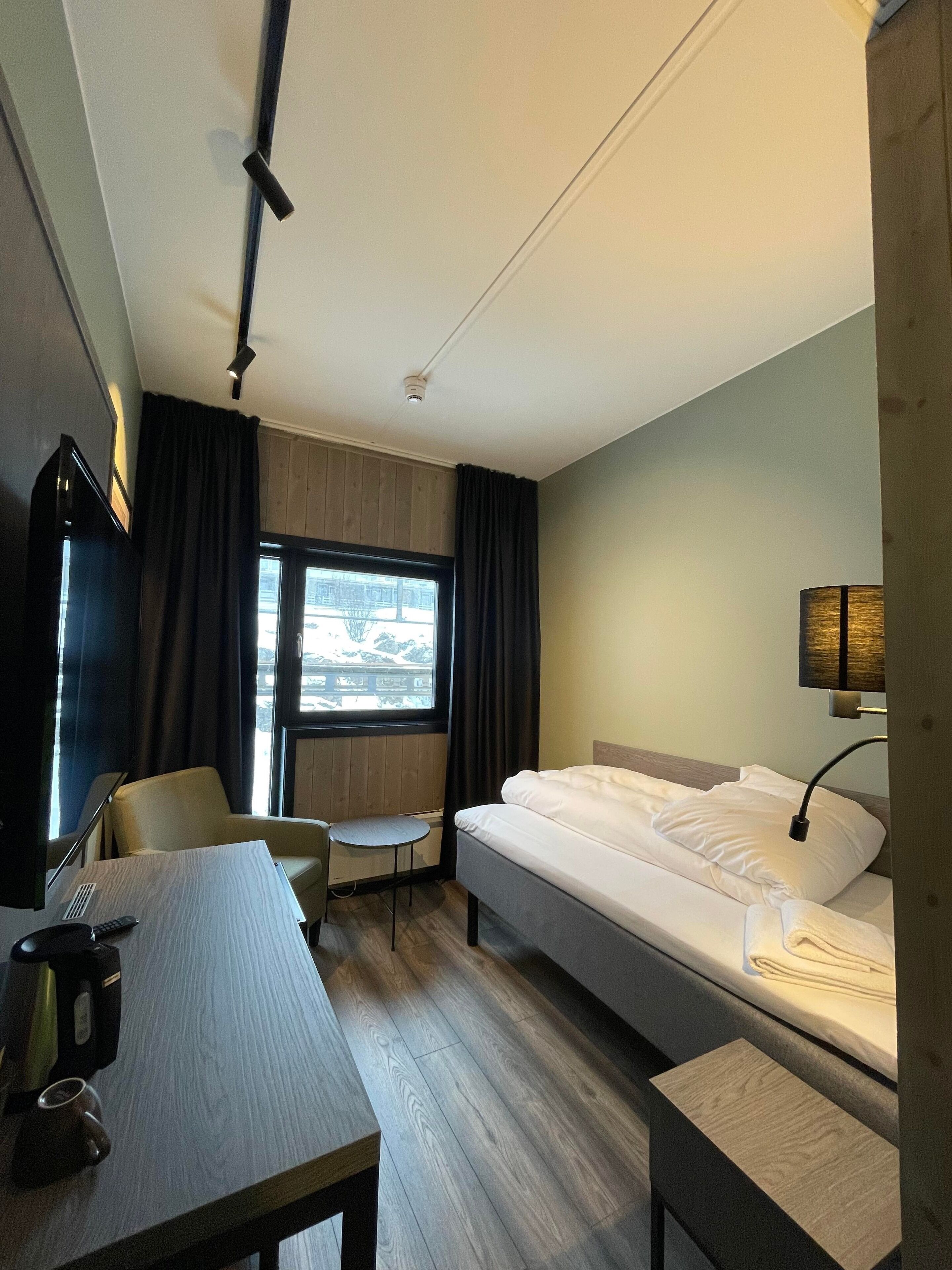 Ein Hotelzimmer mit einem Bett, einem Schreibtisch mit Stuhl, einem kleinen Tisch, einer Lampe und einem Fenster mit Vorhängen.