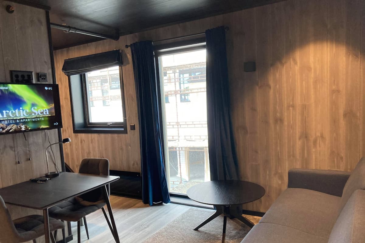 A room with a wooden wall, a TV displaying 'Arctic Sea', a dining table with chairs, a sofa, and a window with curtains.