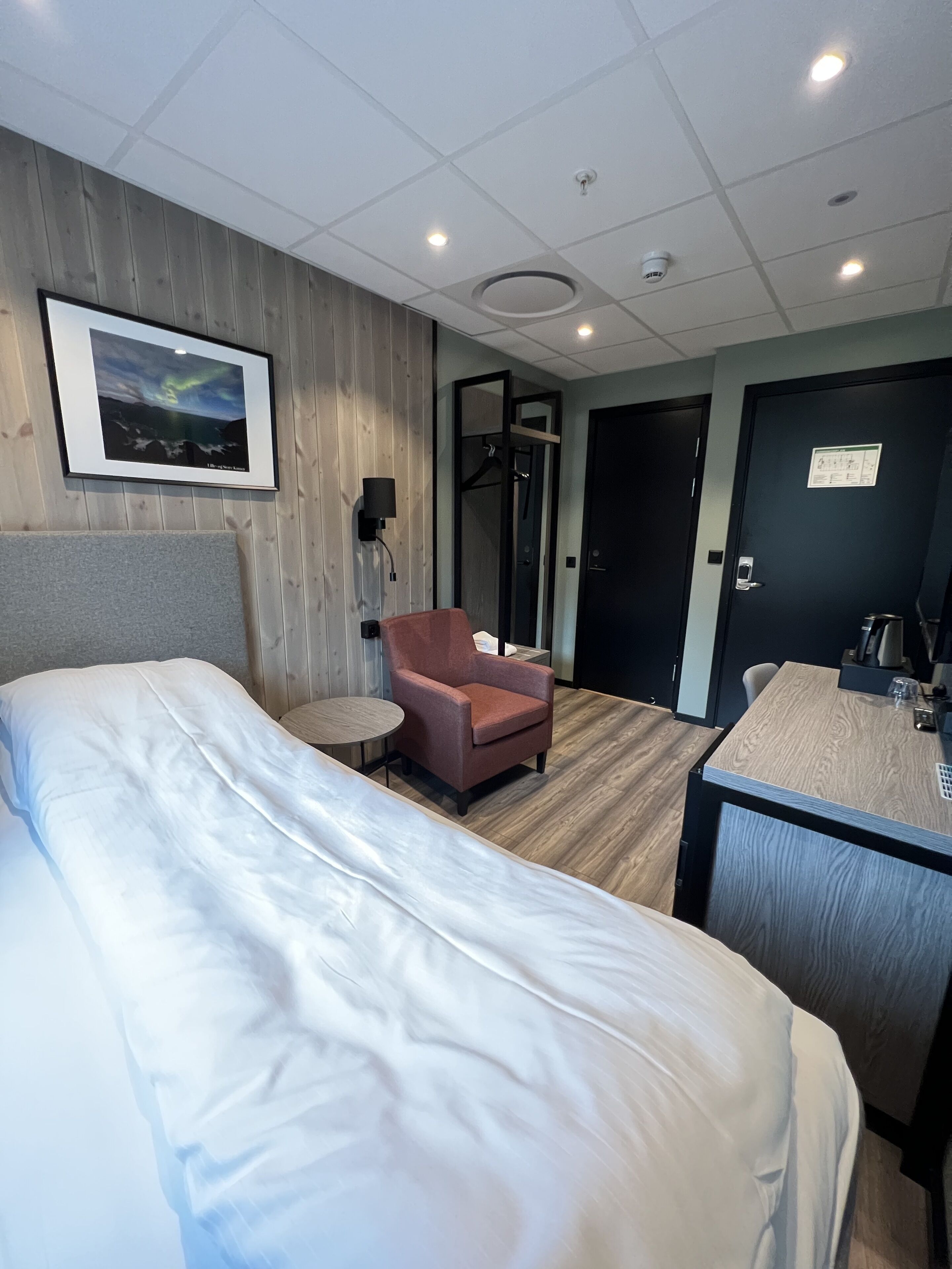 Ett hotellrum med en stor säng, en liten fåtölj, ett skrivbord med en stol, en inramad tavla på väggen och en dörr med en skylt.