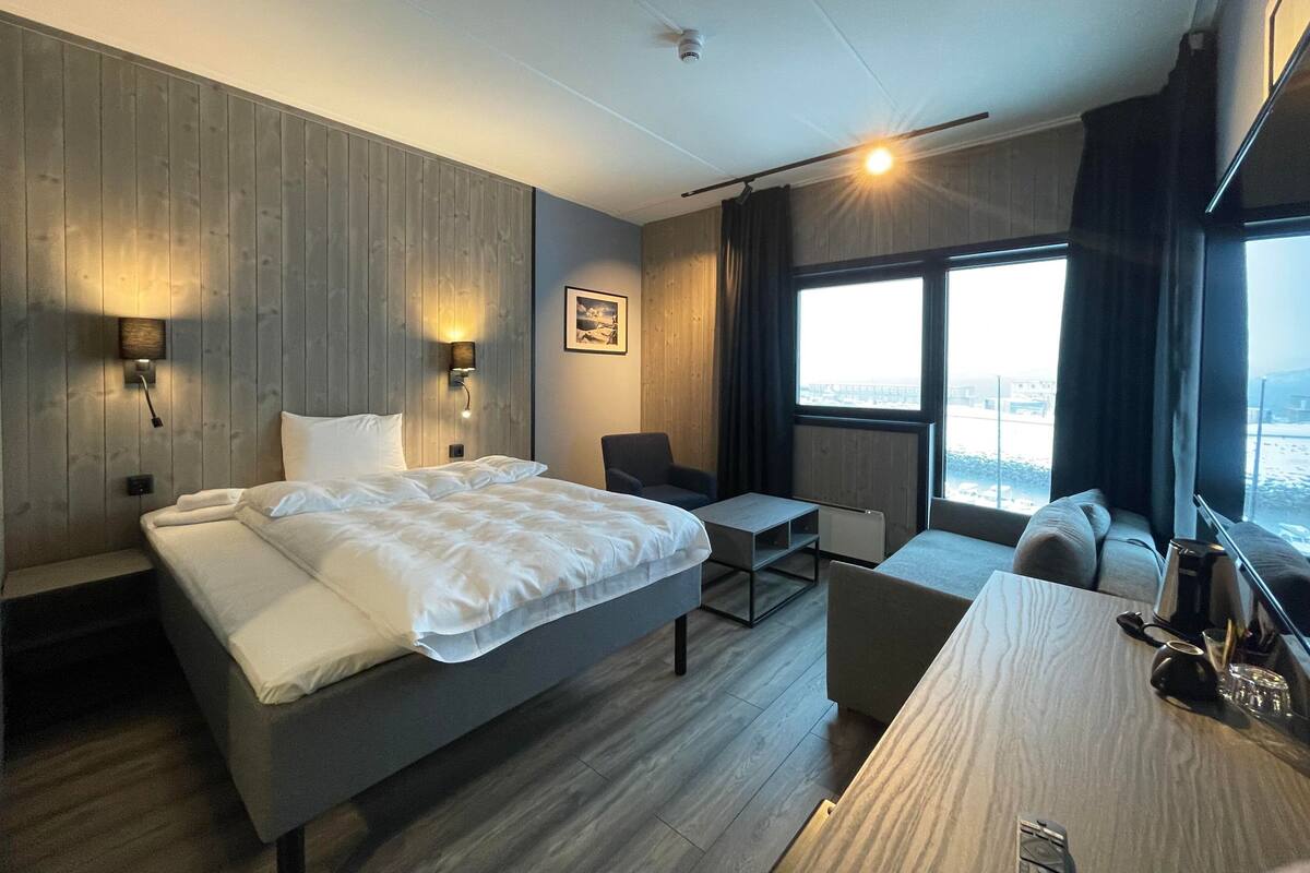 A modern hotel room with a large bed, a desk, a chair, and a view of the city through large windows.