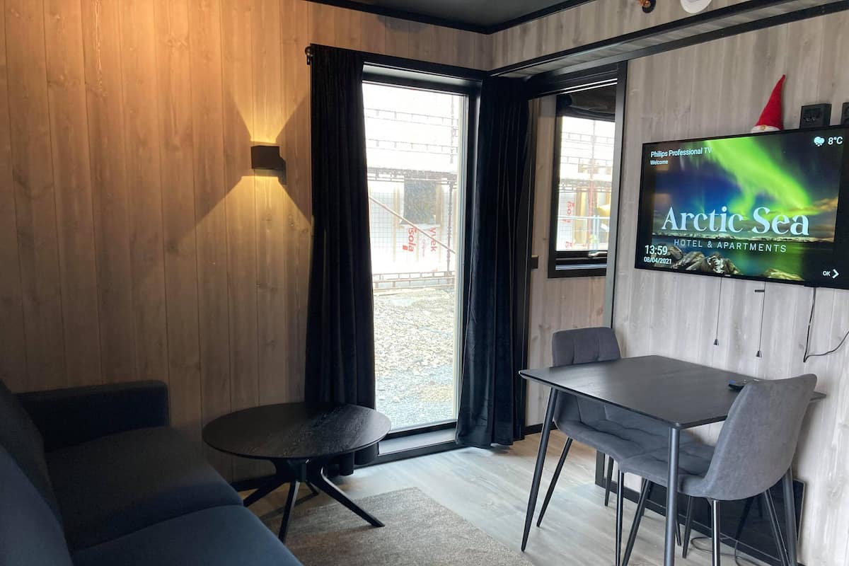 A modern room with a wooden wall, a large TV screen displaying 'Arctic Sea', a sofa, a dining table with chairs, and a window with curtains.