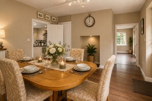A dining area with a round wooden table, beige chairs, a vase with flowers, candles, and a wall clock. - Stubbs House, Loddon, sleeps 20, 2 hot tubs (Loddon)
