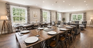 Une salle Ă manger avec une longue table dressĂ©e pour accueillir de nombreux convives, entourĂ©e de chaises, et dotĂ©e de grandes fenĂȘtres et de rideaux.